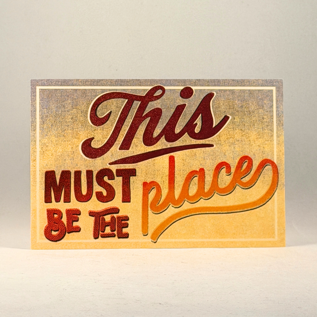 A postcard with the phrase 'This must be the place' in stylized red and orange lettering on a distressed beige-to-yellow background, photographed against a neutral gray background.