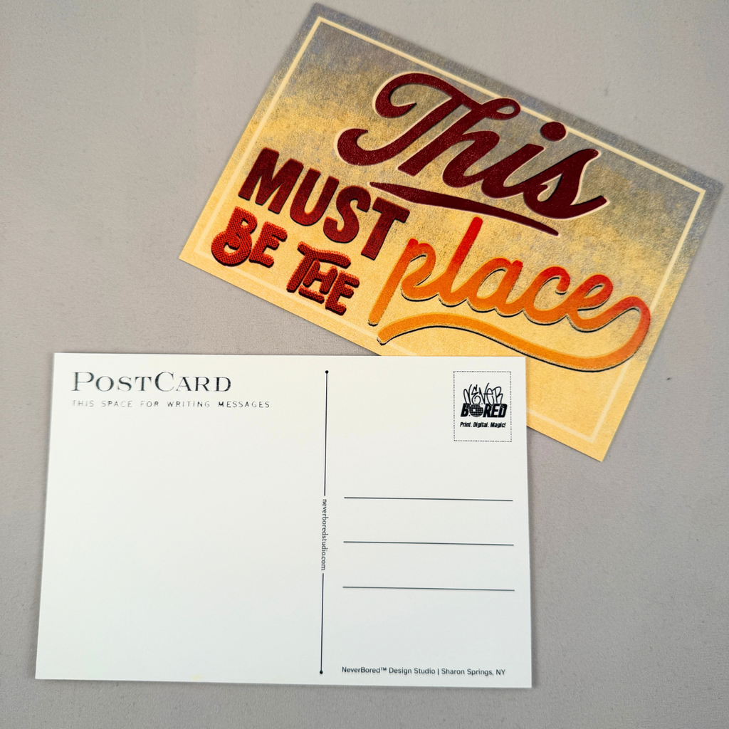 A postcard featuring the front and back on a neutral gray background. The front has a distressed gold background with the phrase 'This MUST BE the place' in stylized red and orange lettering.