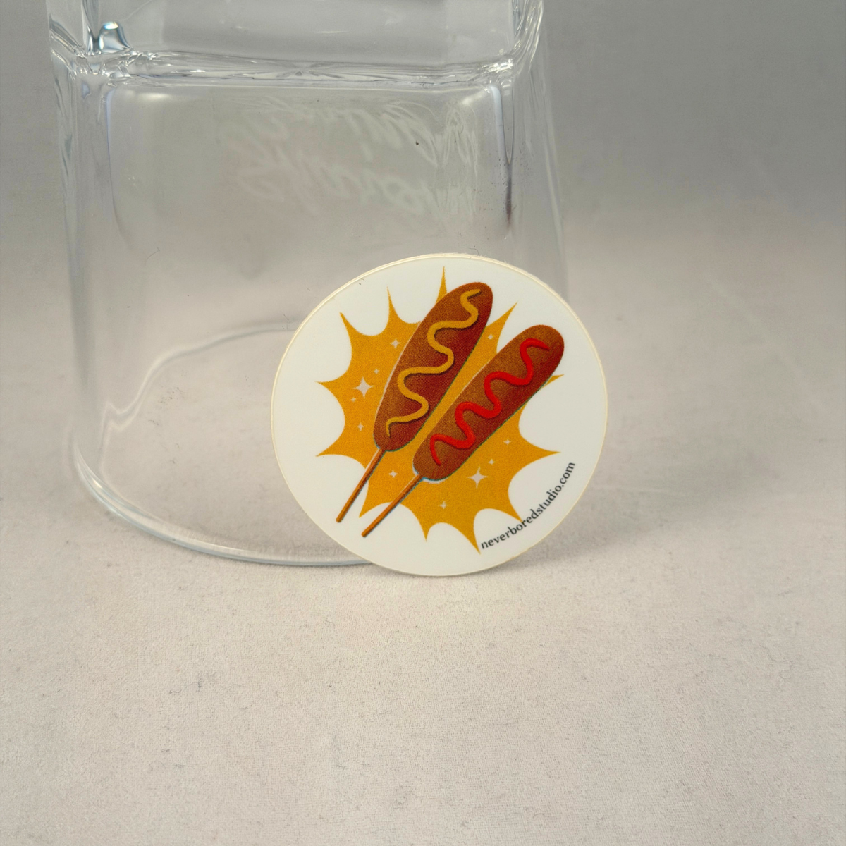 A round white die-cut sticker featuring an illustration of two hot dogs on sticks with mustard and ketchup, placed in front of a clear glass tumbler on a light gray surface.