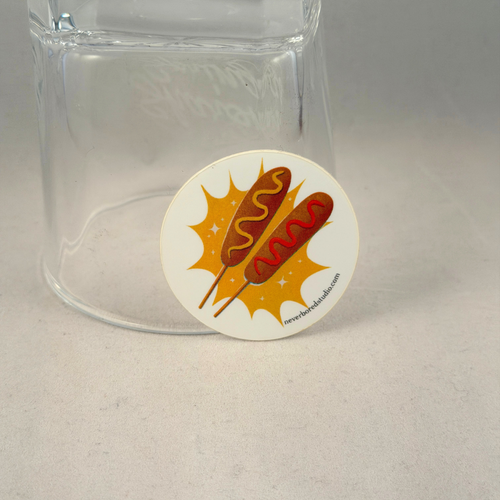 A round white die-cut sticker featuring an illustration of two hot dogs on sticks with mustard and ketchup, placed in front of a clear glass tumbler on a light gray surface.