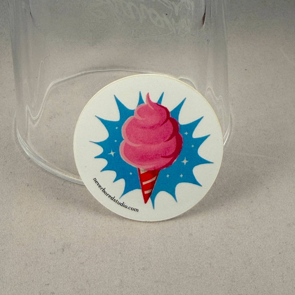 A round white die-cut sticker featuring a pink cotton candy graphic on a blue starburst background, shown resting against a clear glass tumbler on a gray surface.