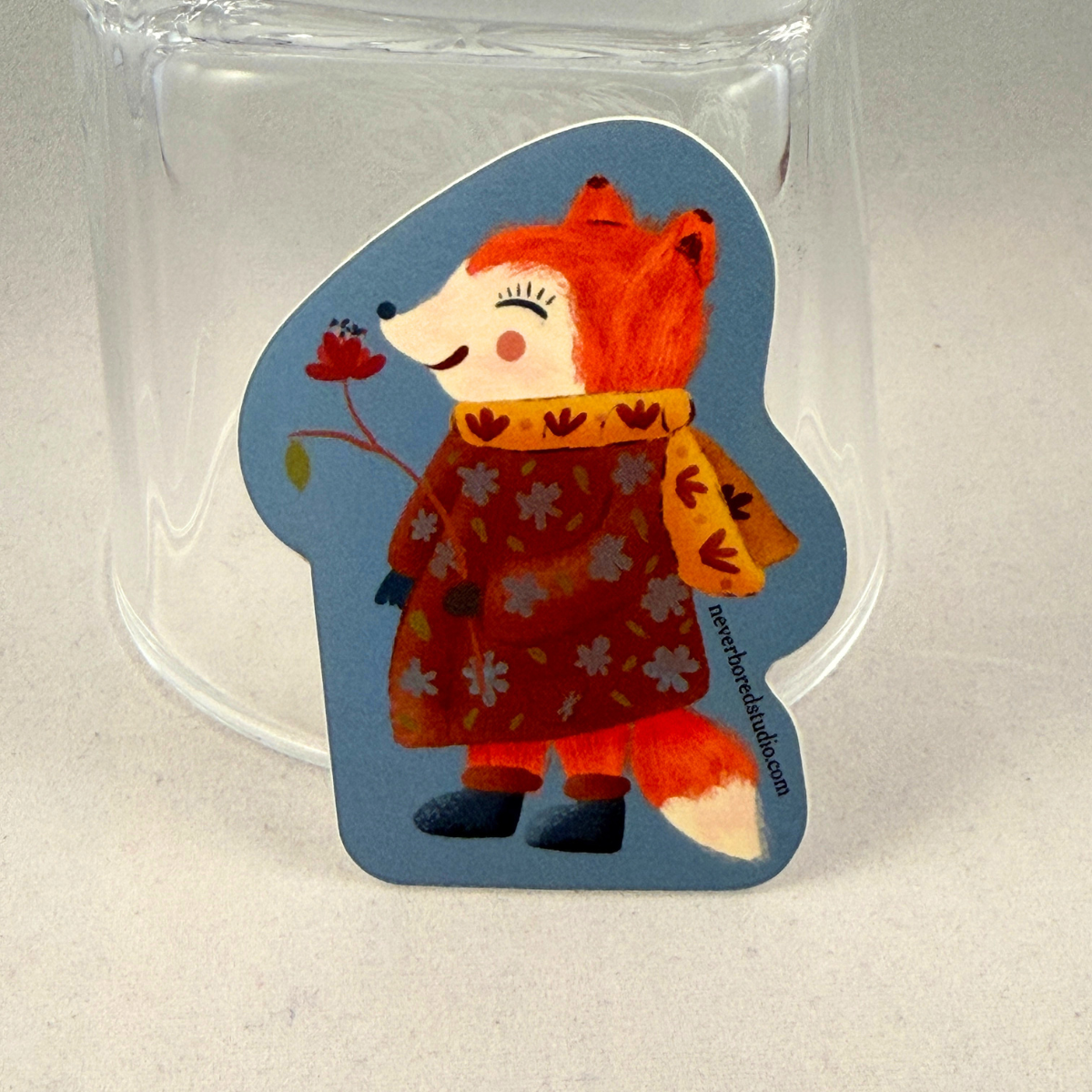 A die-cut sticker featuring an illustration of a smiling orange fox wearing a red floral coat and yellow scarf, set against a blue background. The sticker is displayed upright inside a clear glass tumbler on a light gray surface.