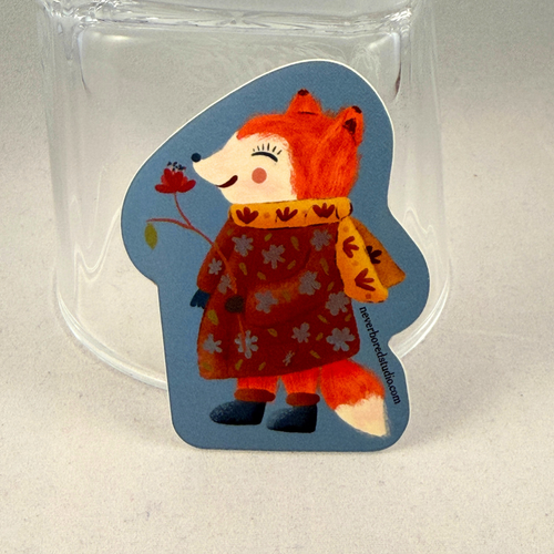 A die-cut sticker featuring an illustration of a smiling orange fox wearing a red floral coat and yellow scarf, set against a blue background. The sticker is displayed upright inside a clear glass tumbler on a light gray surface.