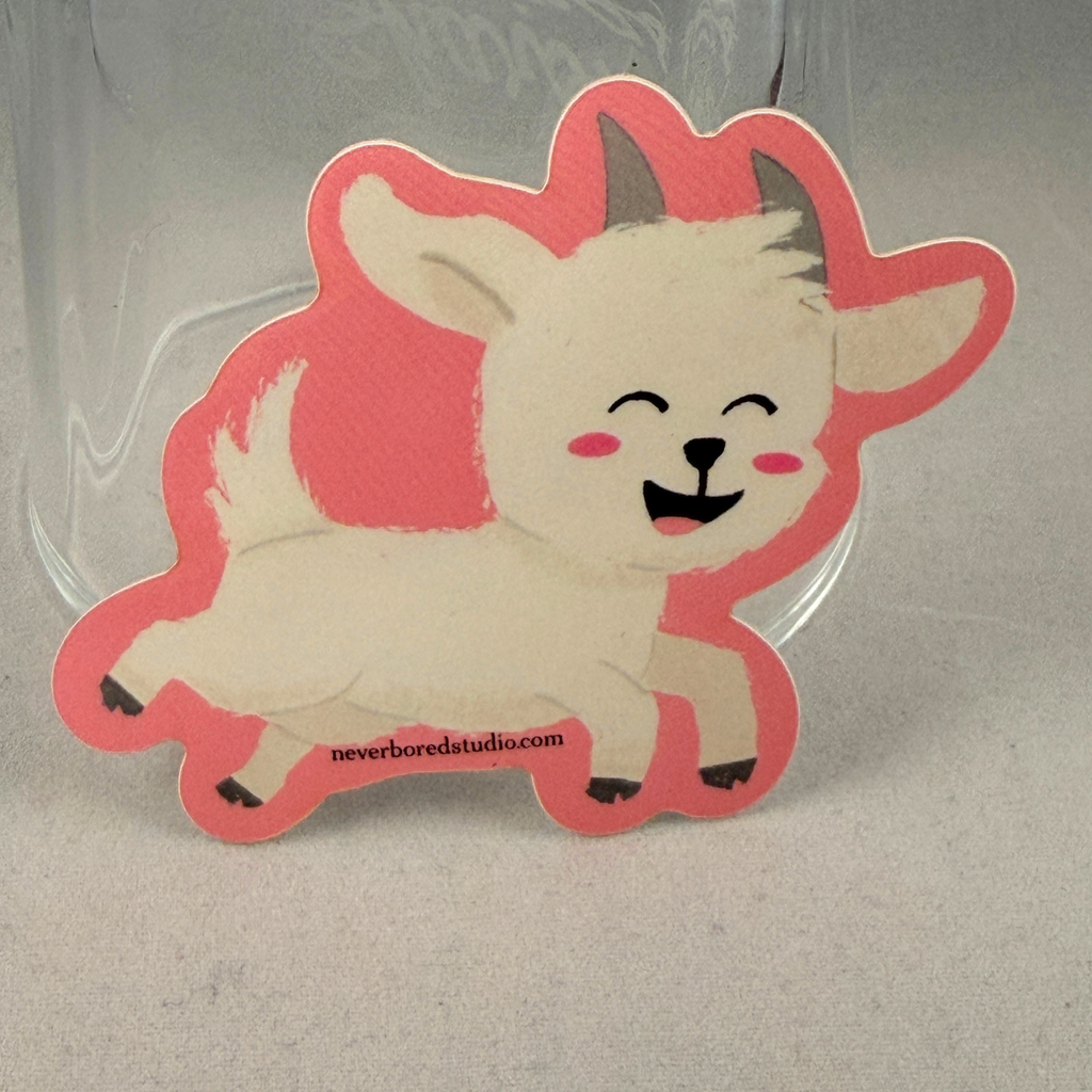 A die-cut sticker featuring a cute cartoon white goat with a pink border, placed on a light gray surface in front of a clear glass.