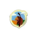 A die-cut sticker featuring an autumn landscape illustration with a large brown and orange leaf, an acorn, green grass, a blue sky, and white clouds, shown on a plain white background.