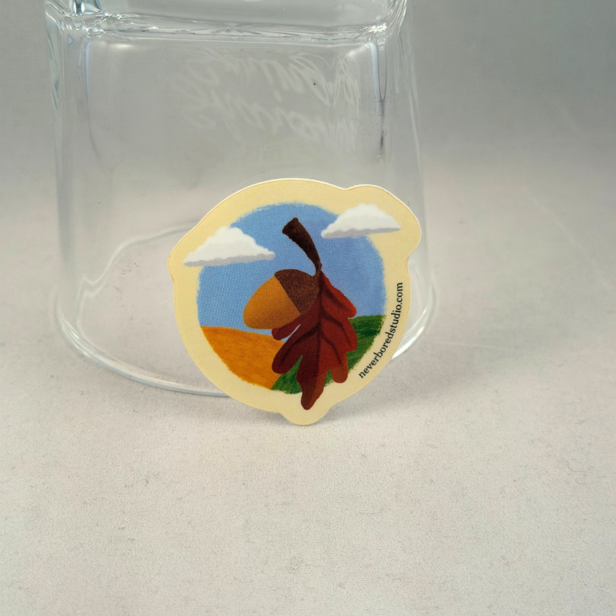 A die-cut sticker featuring an autumn landscape with a brown acorn, red-brown leaves, blue sky, clouds, and grass, shown leaning against a clear glass tumbler on a light gray surface.