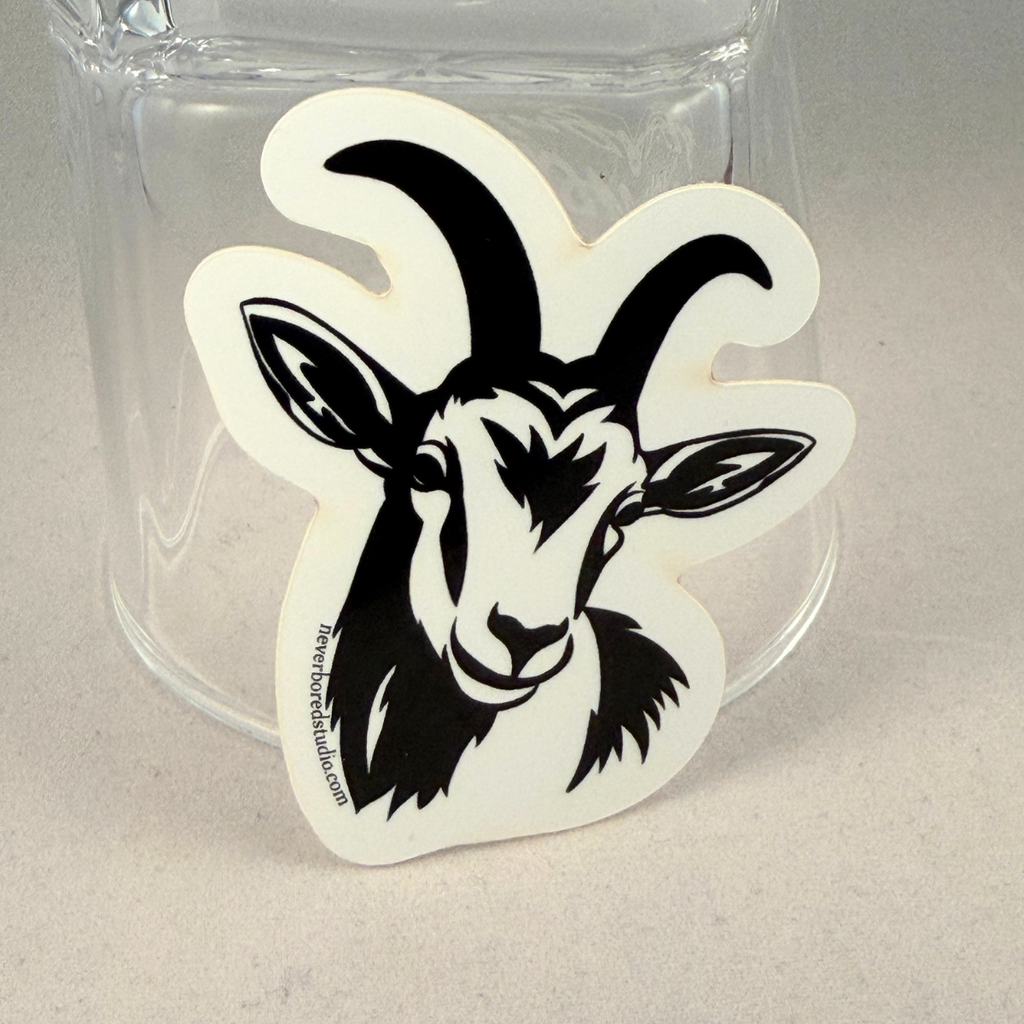 A die-cut sticker featuring a black goat head illustration on a white background, shown leaning against a clear glass tumbler on a light gray surface.