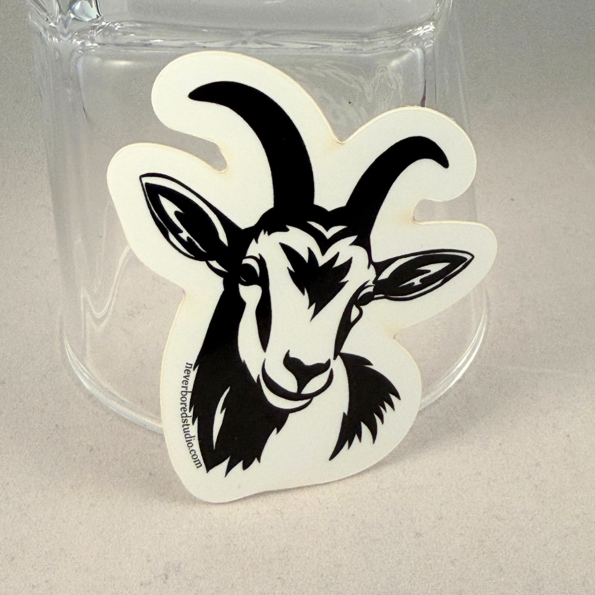 A die-cut sticker featuring a black goat head illustration on a white background, shown leaning against a clear glass tumbler on a light gray surface.