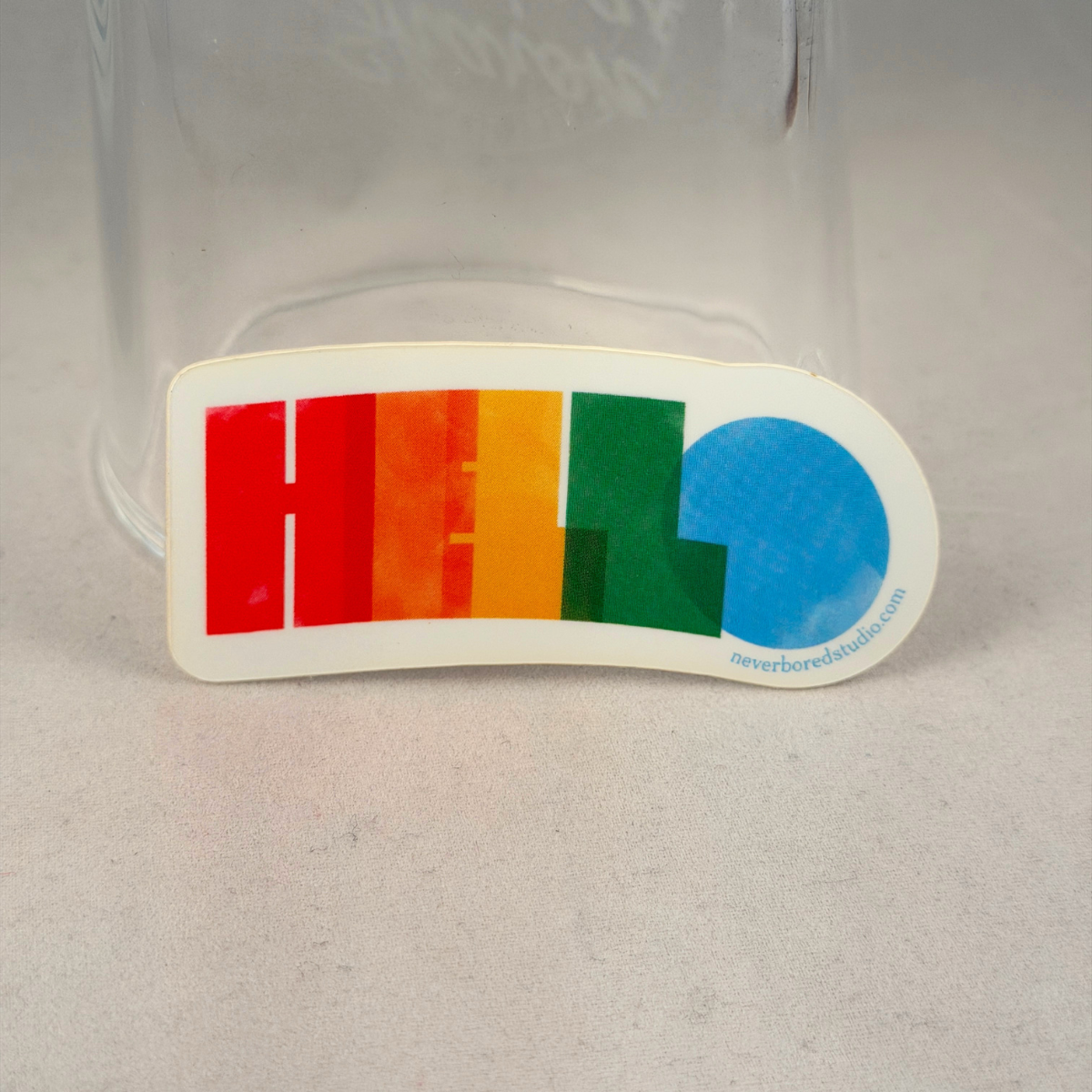 A die-cut sticker featuring the word 'HELLO' in rainbow colors (red, orange, yellow, green, blue) on a white background, placed in front of a clear glass jar on a light gray surface.