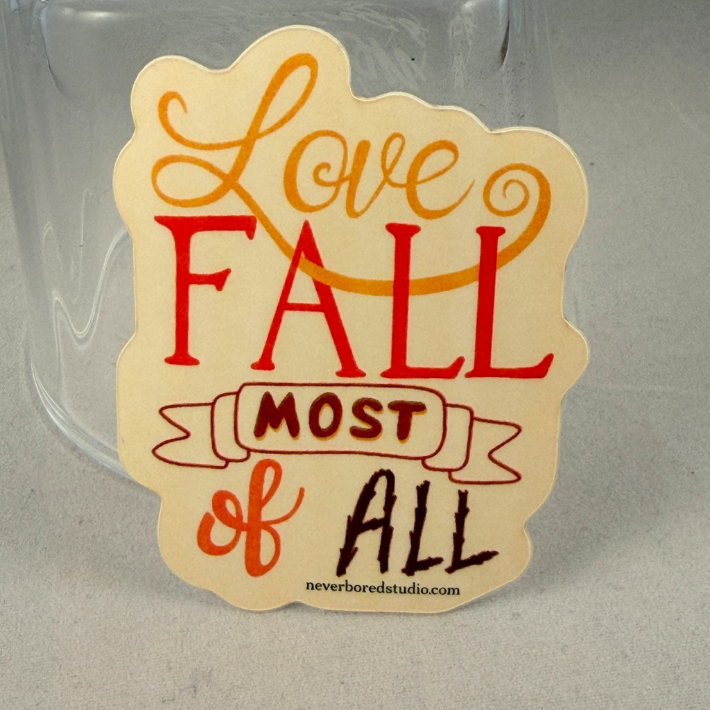 A die-cut sticker with the phrase 'Love FALL most of all' in orange, red, and dark brown lettering on a cream background, shown leaning against a clear glass tumbler on a gray surface.