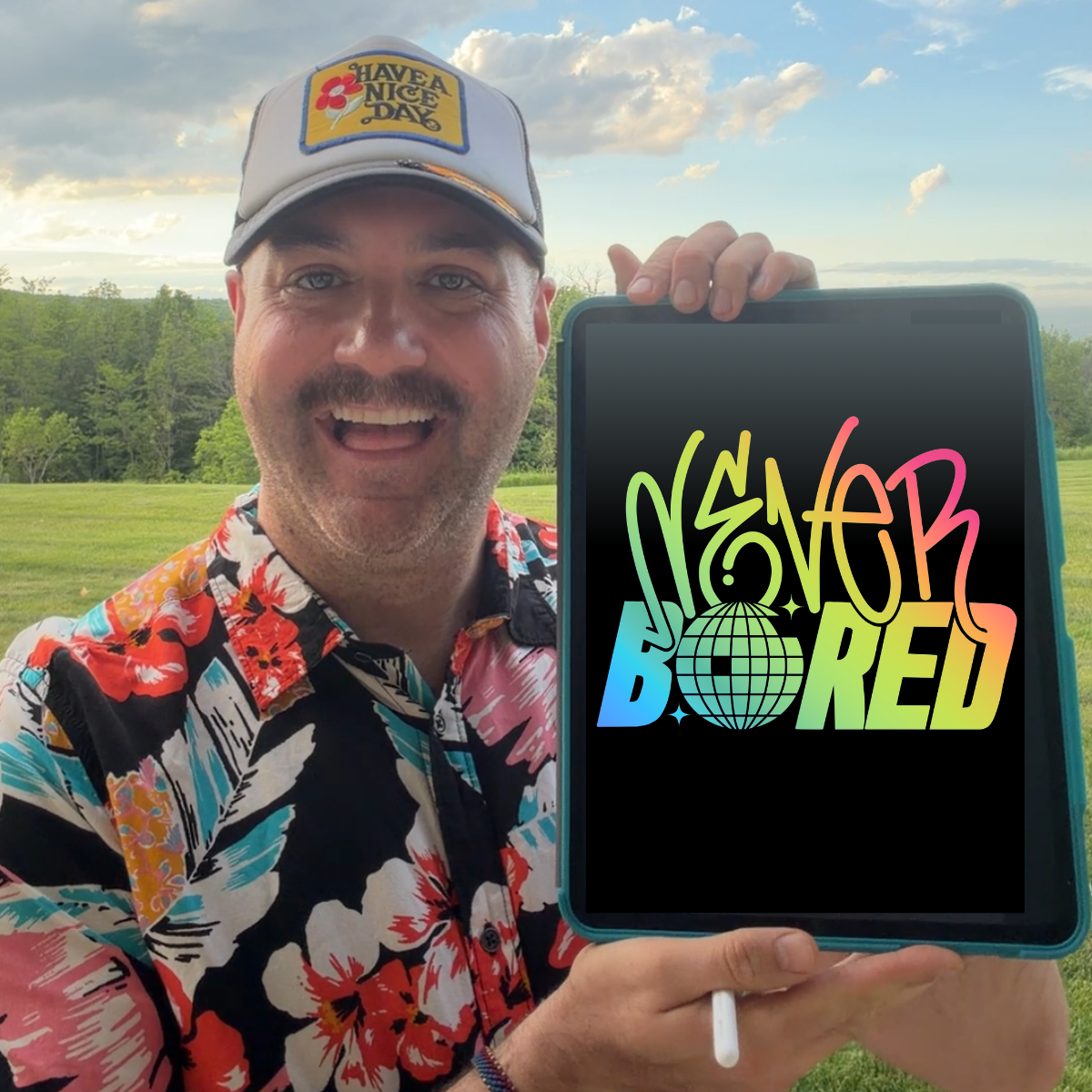 An exuberant man outdoors in a grassy field wearing a colorful floral shirt and a gray 'Have a Nice Day' cap, holding up a tablet that displays the text 'Never Bored' in a rainbow gradient font.